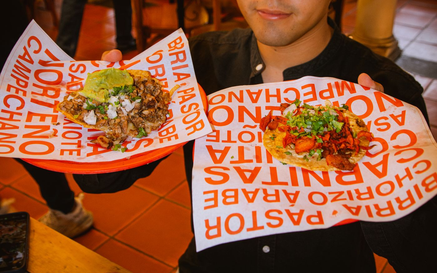 A person holding two paper trays of Mexican street tacos topped with meat, cilantro, and salsa